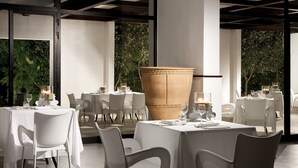 An outdoor dining area with white tables and chairs, a large terracotta pot, and a view of greenery through large windows. - Meditur Puglia by Itafirst Hotels (Carovigno)
