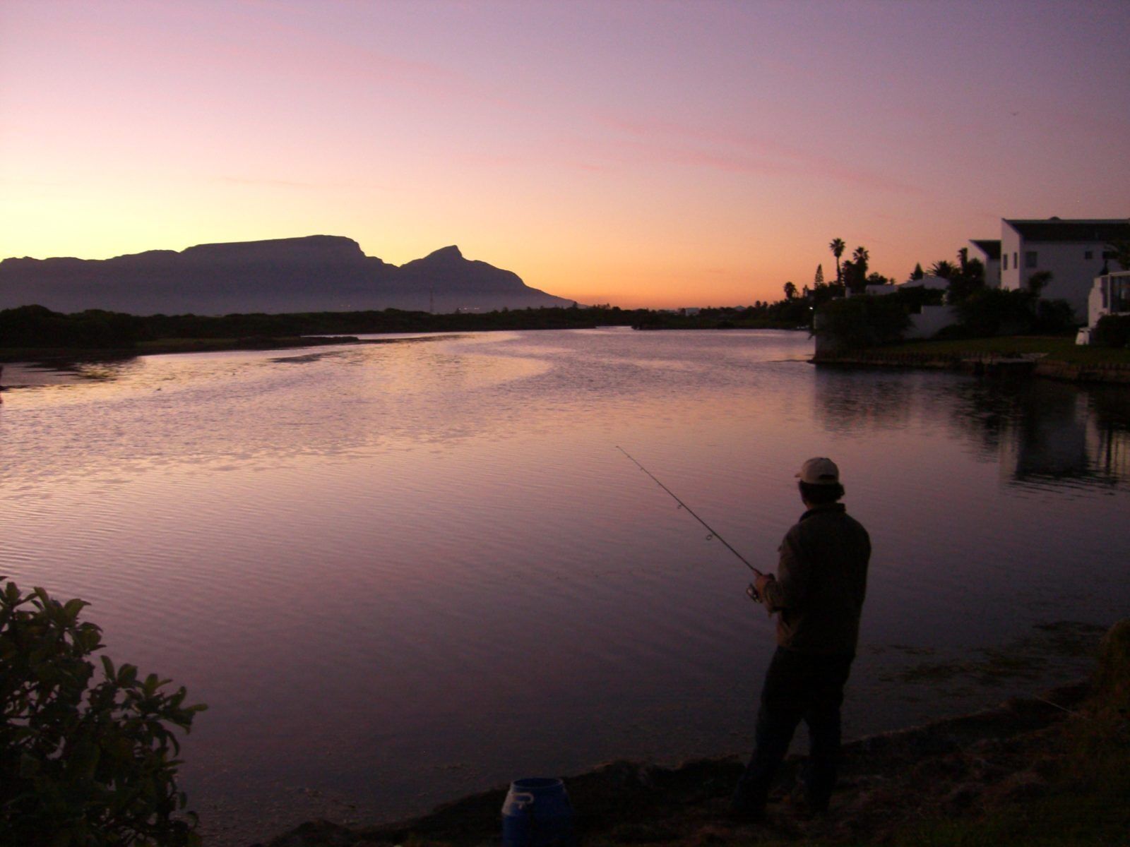 A person fishing by a calm lake at sunset with mountains in the background.