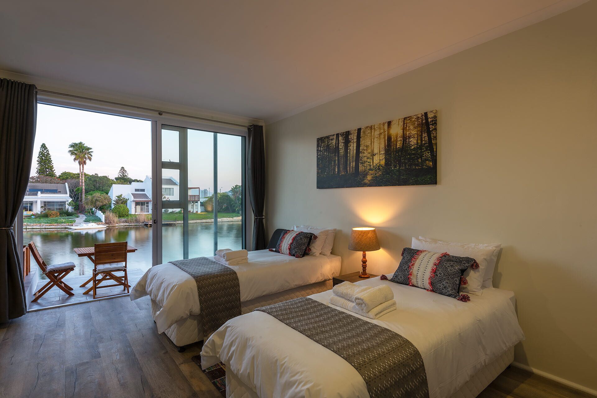 Habitación de hotel con dos camas, un ventanal grande, un cuadro y vistas a un cuerpo de agua y edificios.