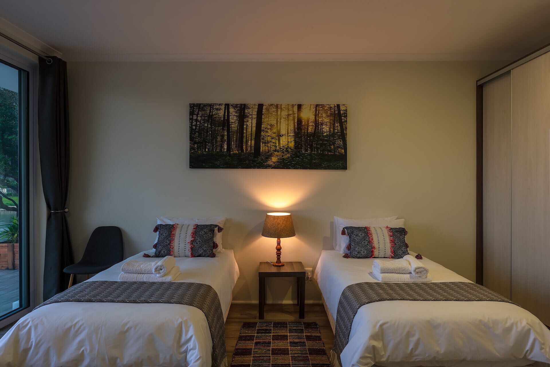 Habitación de hotel con dos camas, un cuadro de un bosque, una lámpara y vistas al exterior.