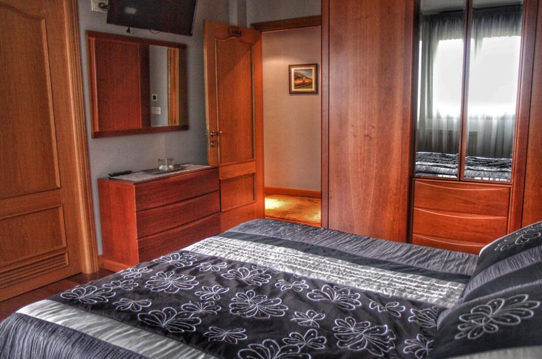 Un dormitorio con cama, armario de madera, cómoda, espejo y ventana con cortinas.
