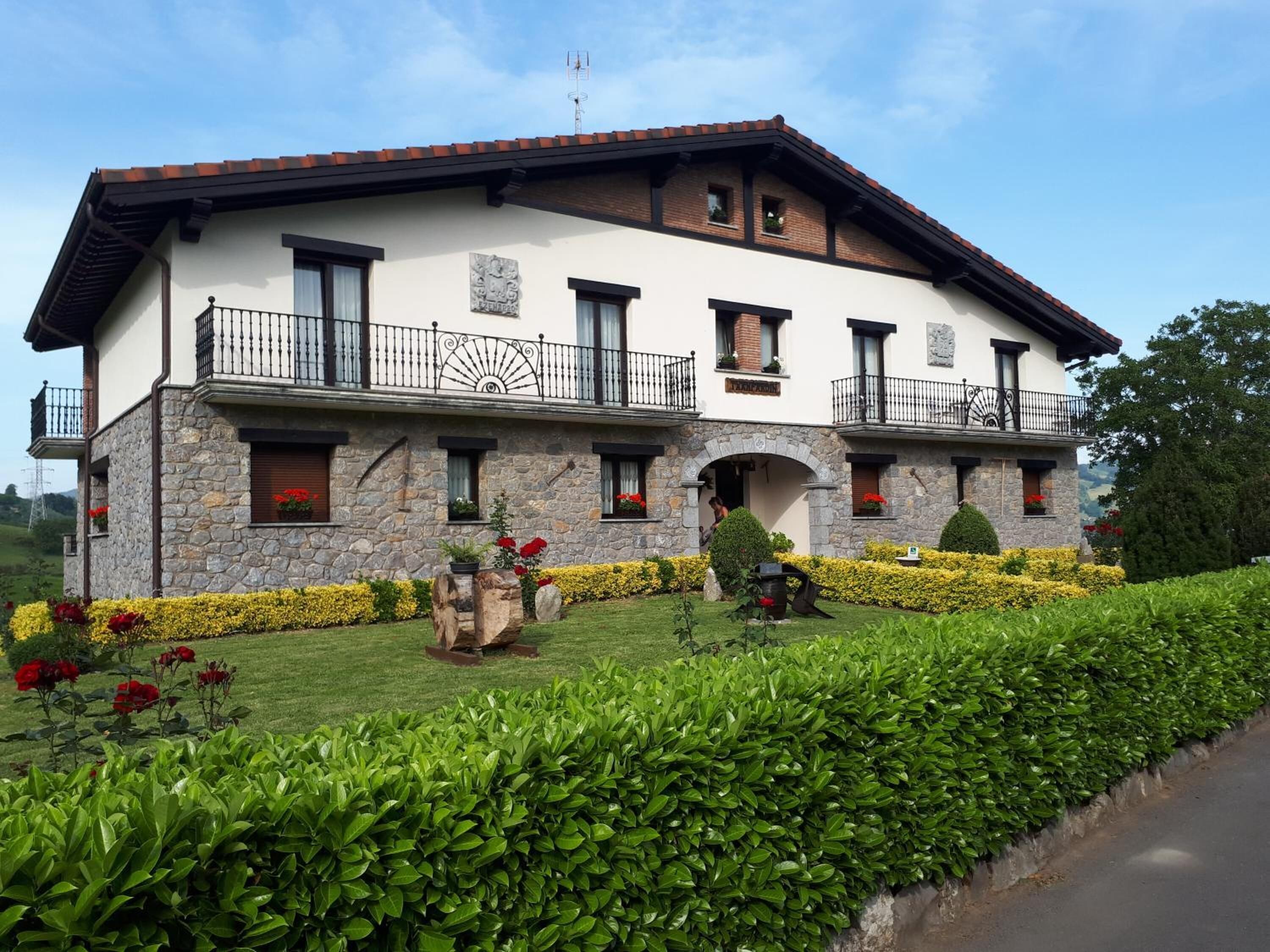 Una casa de dos pisos con paredes exteriores de piedra y yeso, varios balcones con barandillas negras y un jardín bien cuidado con setos y flores.