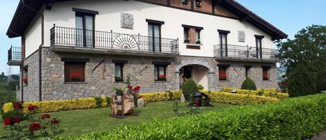 Una casa de dos pisos con paredes exteriores de piedra y yeso, varios balcones con barandillas negras y un jardín bien cuidado con setos y flores.