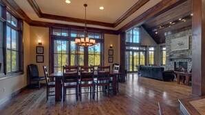 A spacious dining area with a wooden table and chairs, large windows offering a view of the outdoors, and a stone fireplace in the background.