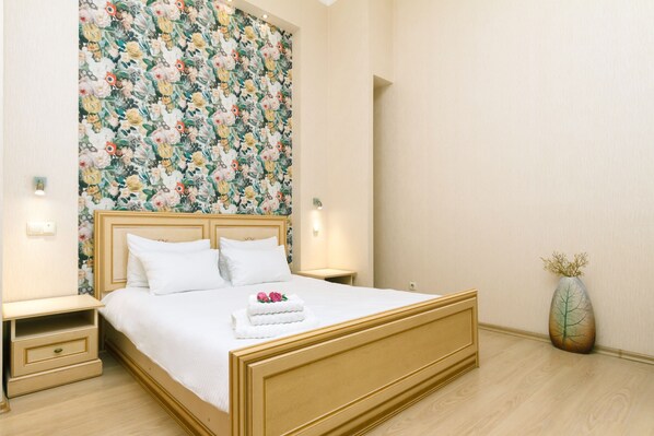 Un lit bien fait, avec du linge de lit blanc et une tête de lit à motifs floraux, une table de chevet avec une lampe et un vase contenant des branches séchées posé sur le sol.