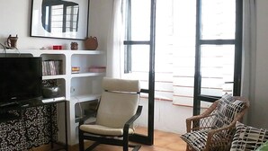 Ein Wohnzimmer mit einer Couch, einem Sessel, einem Fernseher, einem Bücherregal und einem großen Fenster mit Jalousien.