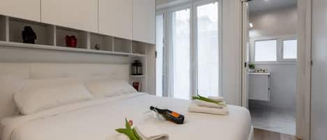 A modern hotel room with a large bed, a minibar, a bathroom visible through an open door, and a view of the outside.