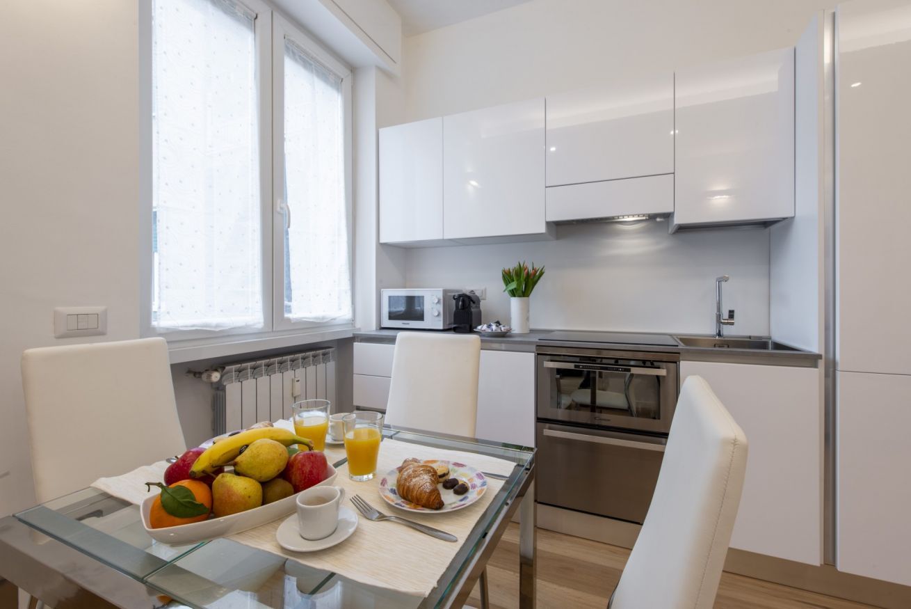 Une cuisine moderne avec des meubles blancs, une table à manger où sont disposés des aliments pour le petit-déjeuner, et une vue sur l’extérieur par une fenêtre.