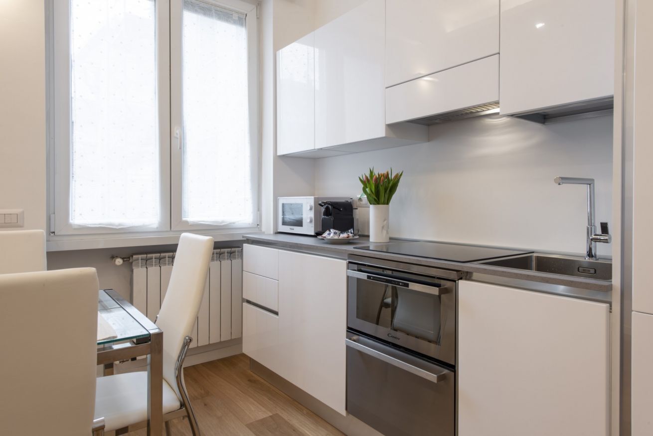 Een moderne keuken met witte kastjes, roestvrijstalen apparatuur, een raam met jaloezieën en een kleine potplant.