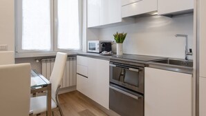 A modern kitchen with white cabinets, stainless steel appliances, a window with blinds, and a small potted plant. - BnButler - Friuli, 24 (Milano)
