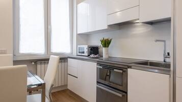 Een moderne keuken met witte kastjes, roestvrijstalen apparatuur, een raam met jaloezieën en een kleine potplant.