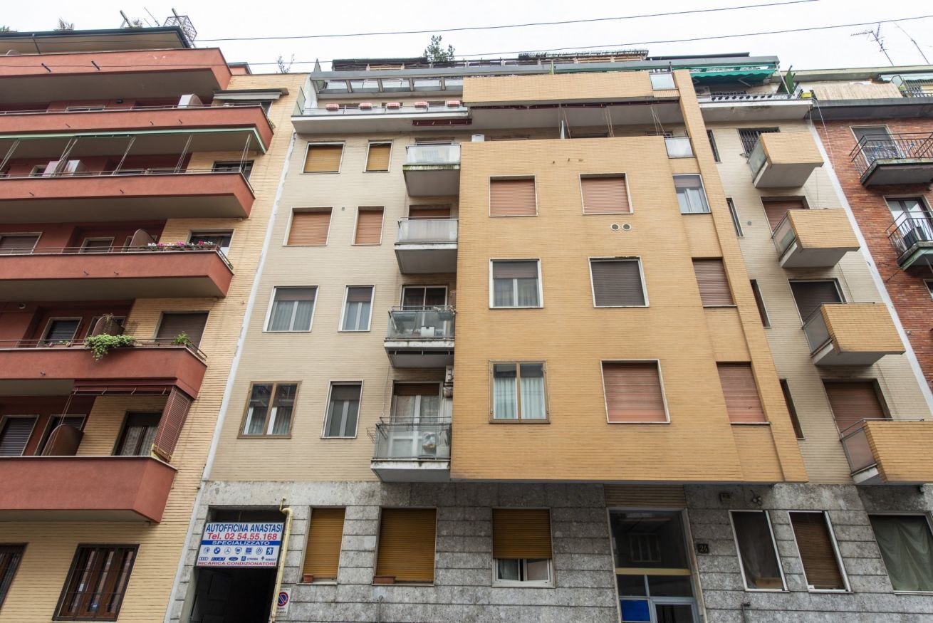 Un edificio residenziale a più piani con balconi, un'attività commerciale al piano terra e un cartello che indica un servizio di autonoleggio.
