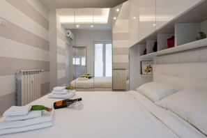 A modern hotel room with a large bed, a bedside table with a bottle and glasses, a radiator, and a window with a view.