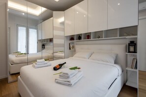 A modern hotel room with a large bed, a minibar, a mirror, and a view of the city.