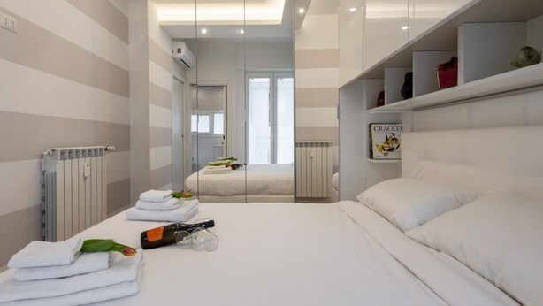 A modern hotel room with a large bed, a bedside table with a bottle and glasses, a radiator, and a window with a view. - BnButler - Friuli, 24 (Milano)