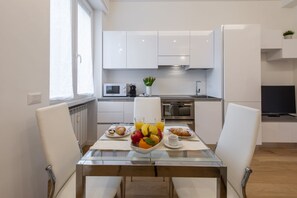 A modern kitchen with white cabinets, a microwave, an oven, and a dining area with a glass table and white chairs. - BnButler - Friuli, 24 (Milano)