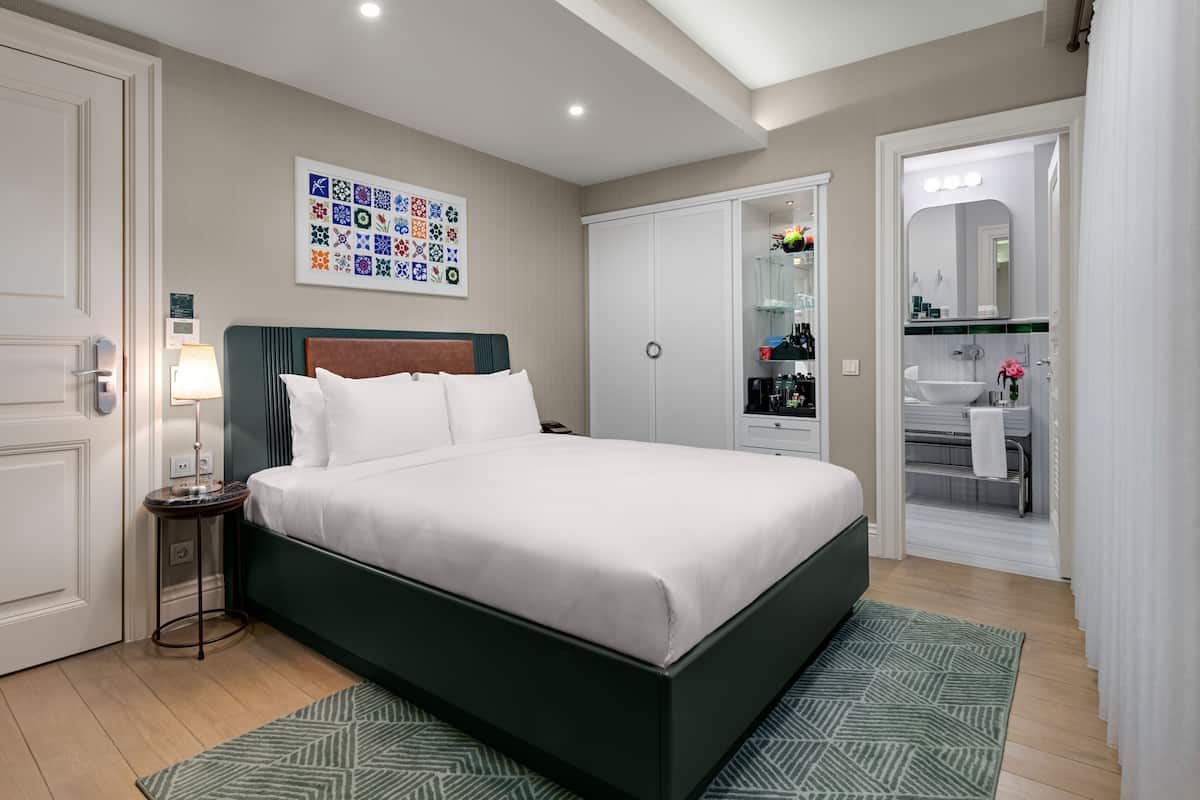 A neatly arranged bedroom with a large bed, a bedside table with a lamp, a wardrobe, and a bathroom visible through an open door.