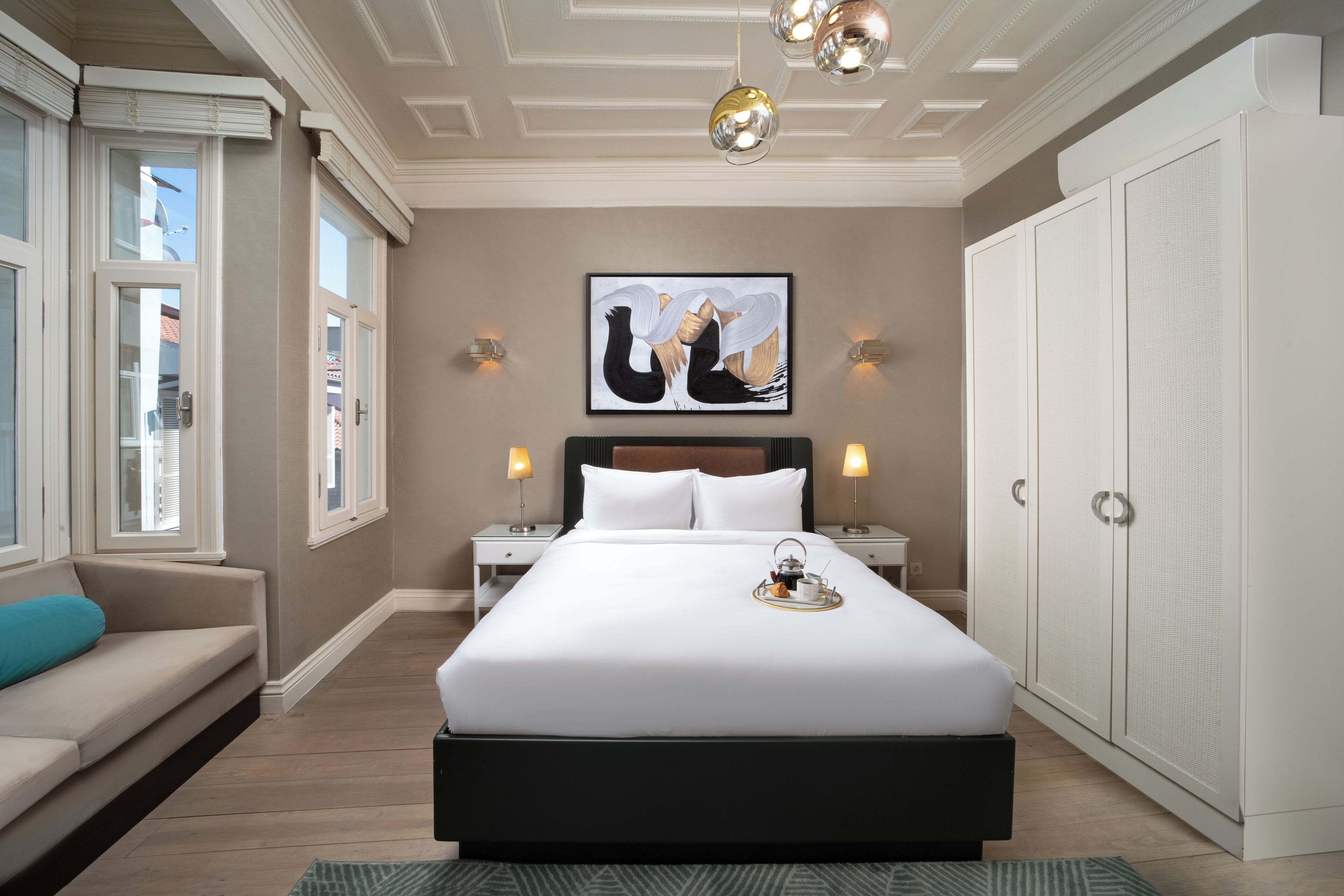 A hotel room with a large bed, two bedside tables, a nightstand, a sofa, a large wardrobe, and a painting on the wall.