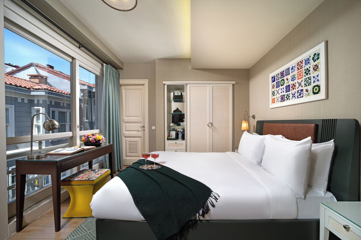 A hotel room with a large bed, a desk with a lamp, a window with curtains, a framed picture on the wall, and a closet.