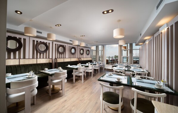 A modern restaurant with a long counter, white chairs, and a high ceiling with recessed lighting.
