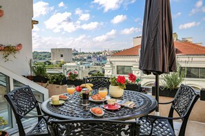 Une table ronde en métal, surmontée d’un parasol noir, entourée de chaises, sur un balcon agrémenté de plantes en pot et offrant une vue sur la ville.