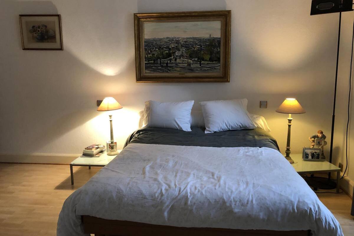 Un lit bien fait, avec une literie blanche, deux lampes de chevet, un tableau représentant un paysage urbain encadré au mur, et une petite table sur laquelle se trouvent un téléphone et des livres.