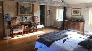 A rustic room with exposed wooden beams, a bed with a dark headboard, a desk with a chair, a radiator, and a window with curtains. - Chambres d'Hotes Las Caneres (Castelnau-d'Arbieu)
