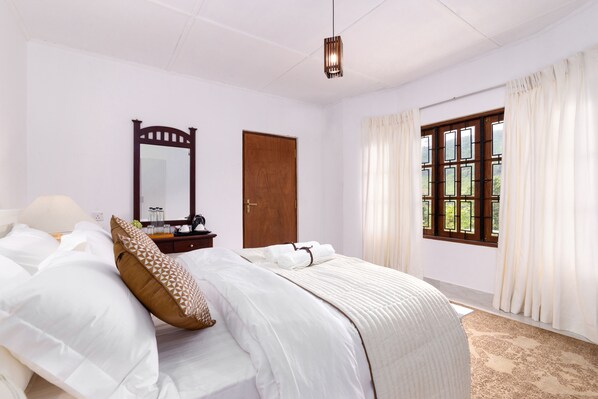 A neatly made bed with white linens and a patterned pillow, a wooden door, a mirror, and a window with curtains.