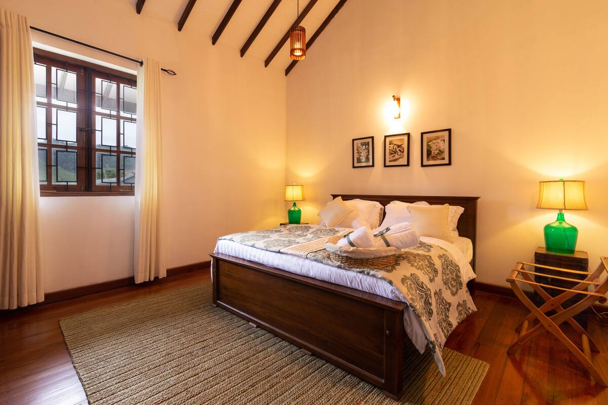 A bedroom with a wooden bed, a window with curtains, a green lamp, and framed pictures on the wall.