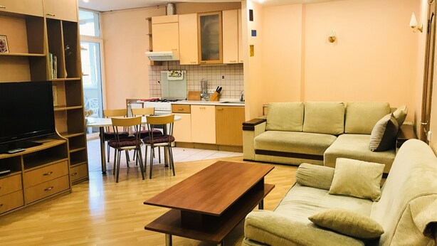 Un salon moderne avec une table basse en bois, un canapé beige et un téléviseur posé sur un meuble en bois. On aperçoit une cuisine en arrière-plan, avec une cuisinière et des placards.