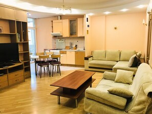 Ein modernes Wohnzimmer mit Couchtisch aus Holz, beiger Couch und einem Fernseher auf einem Holzschrank. Im Hintergrund ist eine Küche mit Herd und Schränken zu sehen.