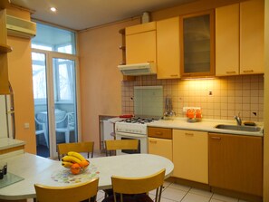 Eine kompakte Küche mit Gasherd, Spüle und Holzschränken. Ein Esstisch mit Stühlen und eine Schüssel mit Obst sind ebenfalls vorhanden.