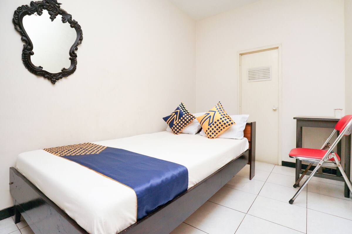 A single bed with a white and blue bedspread, two patterned pillows, a red chair, and a mirror on the wall.