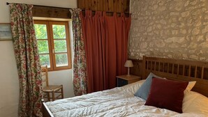 A bedroom with a wooden bed, a nightstand with a lamp, a chair, and a window with curtains. - Renovated farmhouse with stunning views & heated pool (Verteillac)