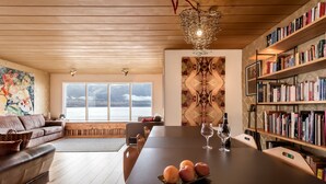 A modern living room with a wooden ceiling, a large window, a sofa, a dining table with fruit and wine glasses, and a bookshelf. - The Crib Rocks (Borgarnes)