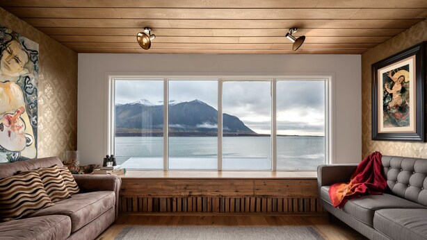 A living room with a large window overlooking a lake and mountains, two sofas, a wooden floor, and a painting on the wall. - The Crib Rocks (Borgarnes)