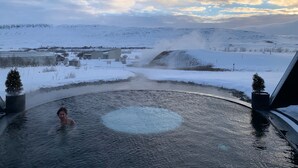 A person in a hot spring surrounded by a snowy landscape with a view of distant hills and a cloudy sky. - The Crib Rocks (Borgarnes)