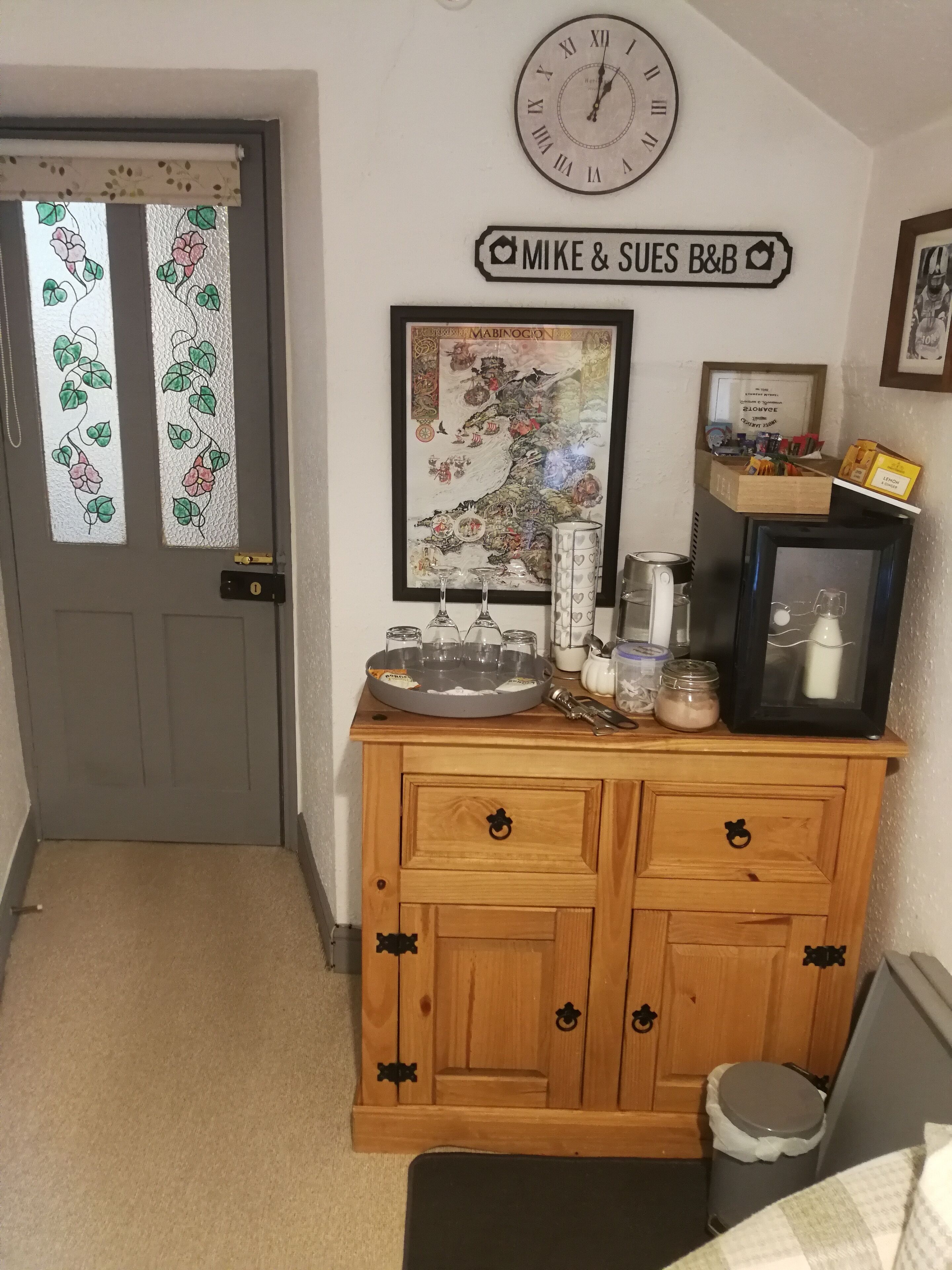 A small, cozy room with a wooden cabinet, a clock, a framed map, and a sign reading "Mike & Sue's B&B".
