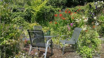 An outdoor seating area with metal chairs and a glass table surrounded by lush greenery and various flowering plants.
