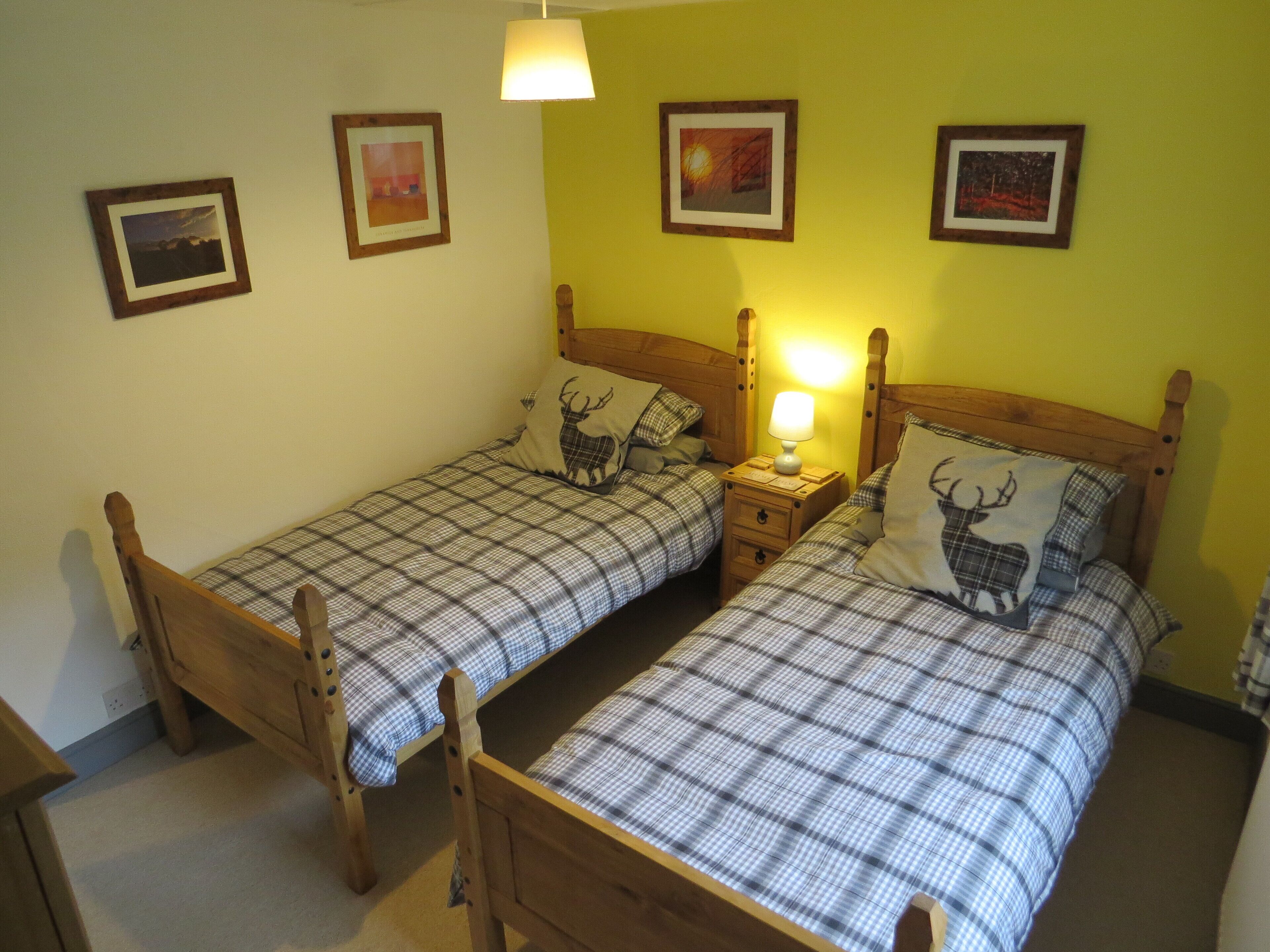 Una habitación con dos camas de madera, cada una con ropa de cama de cuadros y una almohada con forma de ciervo. Hay tres cuadros enmarcados en la pared, una mesita de noche con una lámpara y un cabecero de madera.