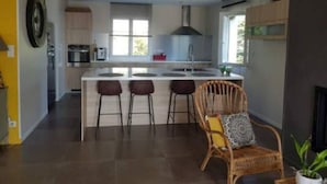 A modern kitchen with a central island, bar stools, and a wicker chair. - Villa "Chjosella" (Santa Reparata di Balagna)