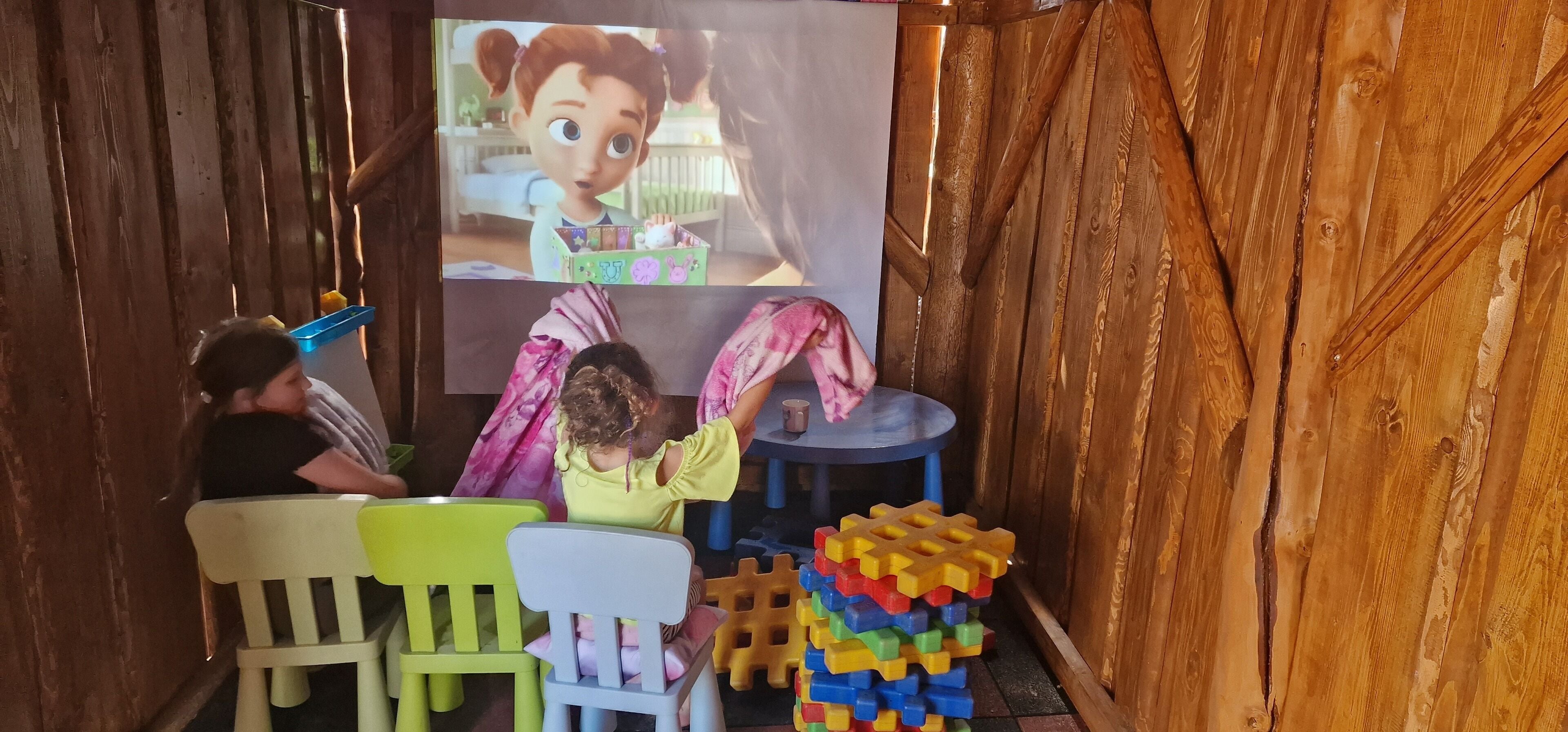 A room with wooden walls, a projector screen displaying an animated character, two children playing with toys, and a small table with a cup.