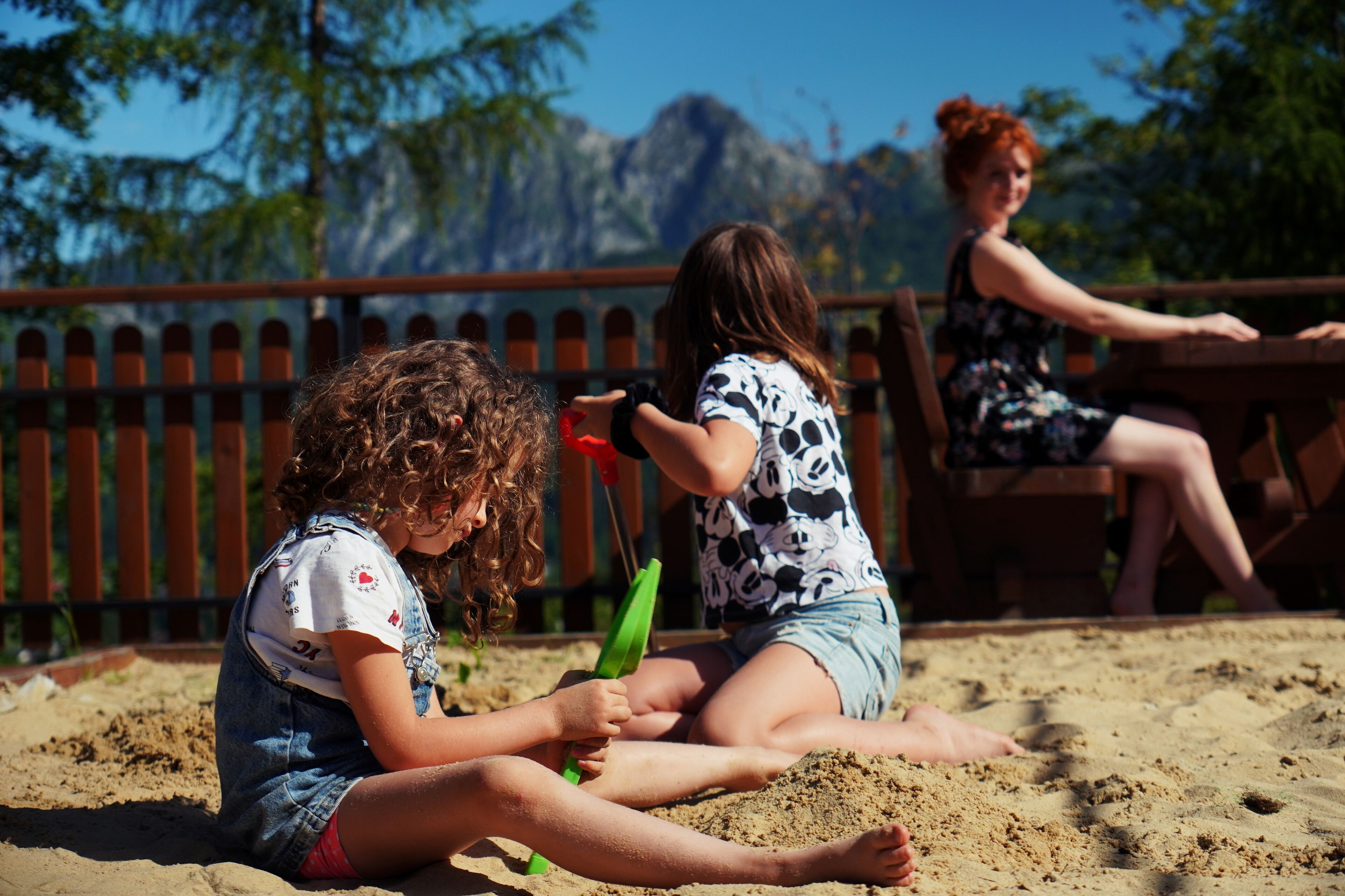 Children playing in sand with a wooden fence and trees in the background.