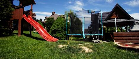 A playground with a red slide, a trampoline, and a wooden playhouse.