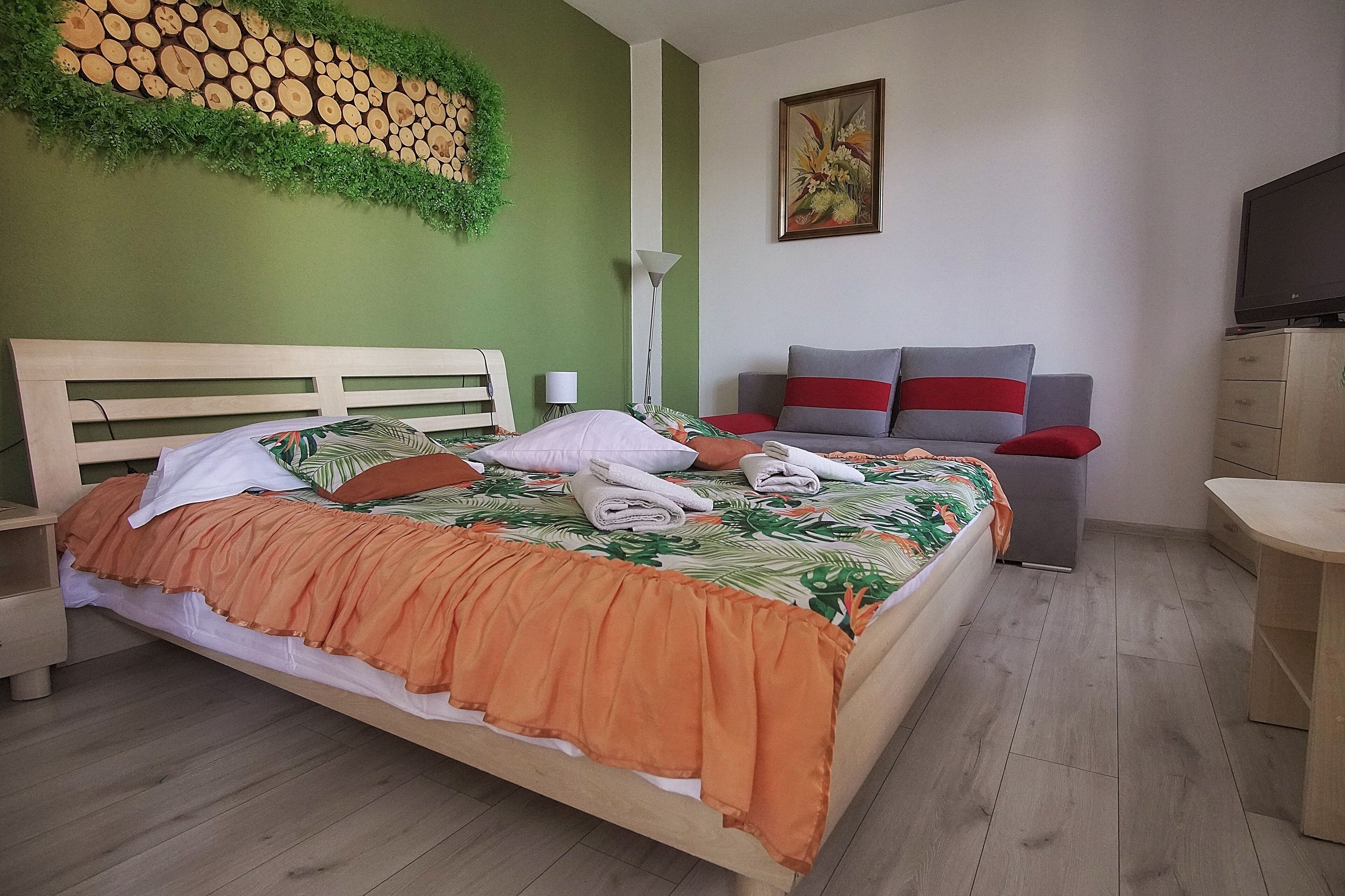 Ein ordentlich bezogenes Bett mit einem tropischen Bettbezug, ein Nachttisch mit einer Lampe und eine Wand mit einem Holzdekor und Grünpflanzen.