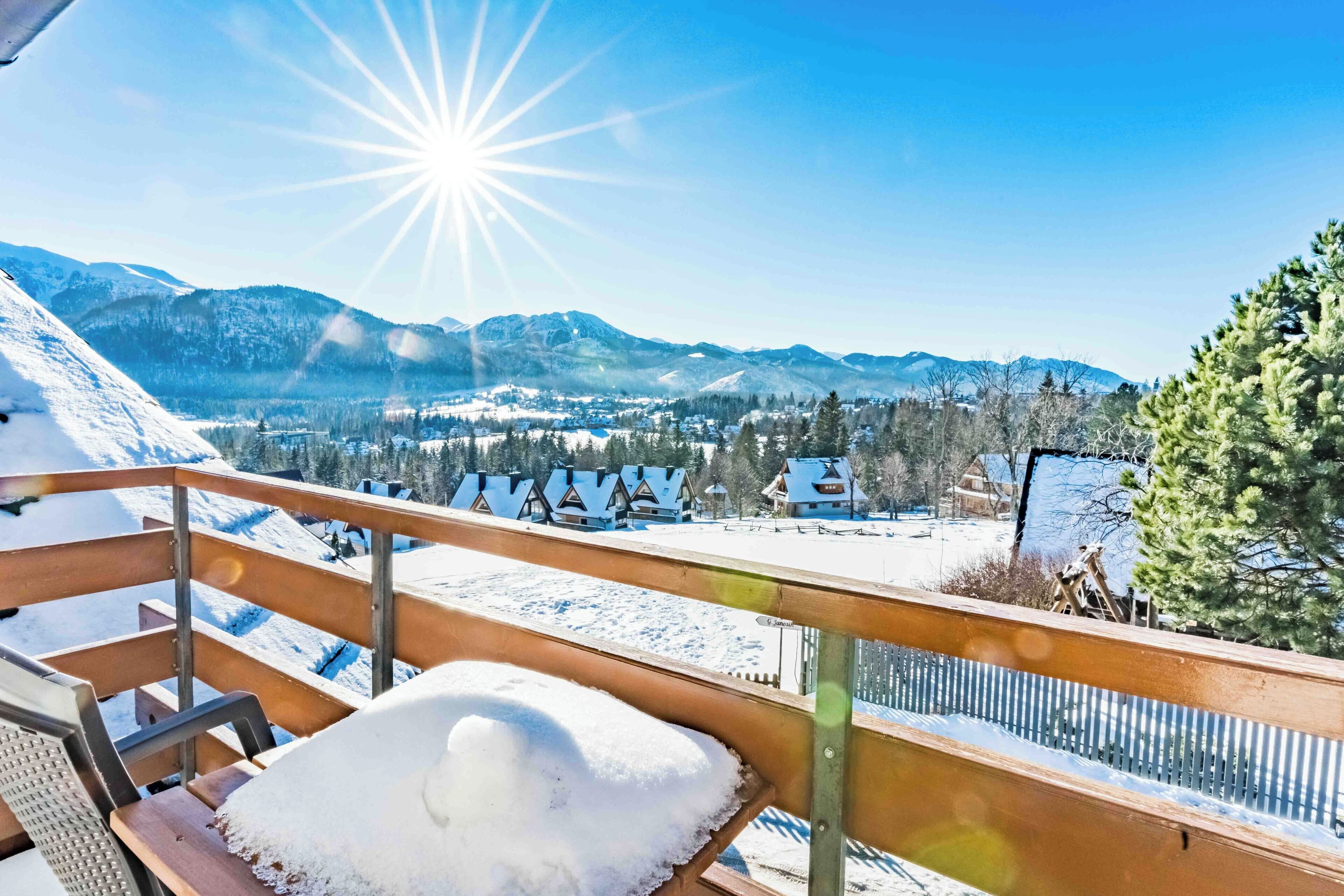 Ein mit Schnee bedeckter Balkon mit einer hölzernen Bank und einem Metallgeländer mit Blick auf eine gebirgige Landschaft mit Häusern und Bäumen.