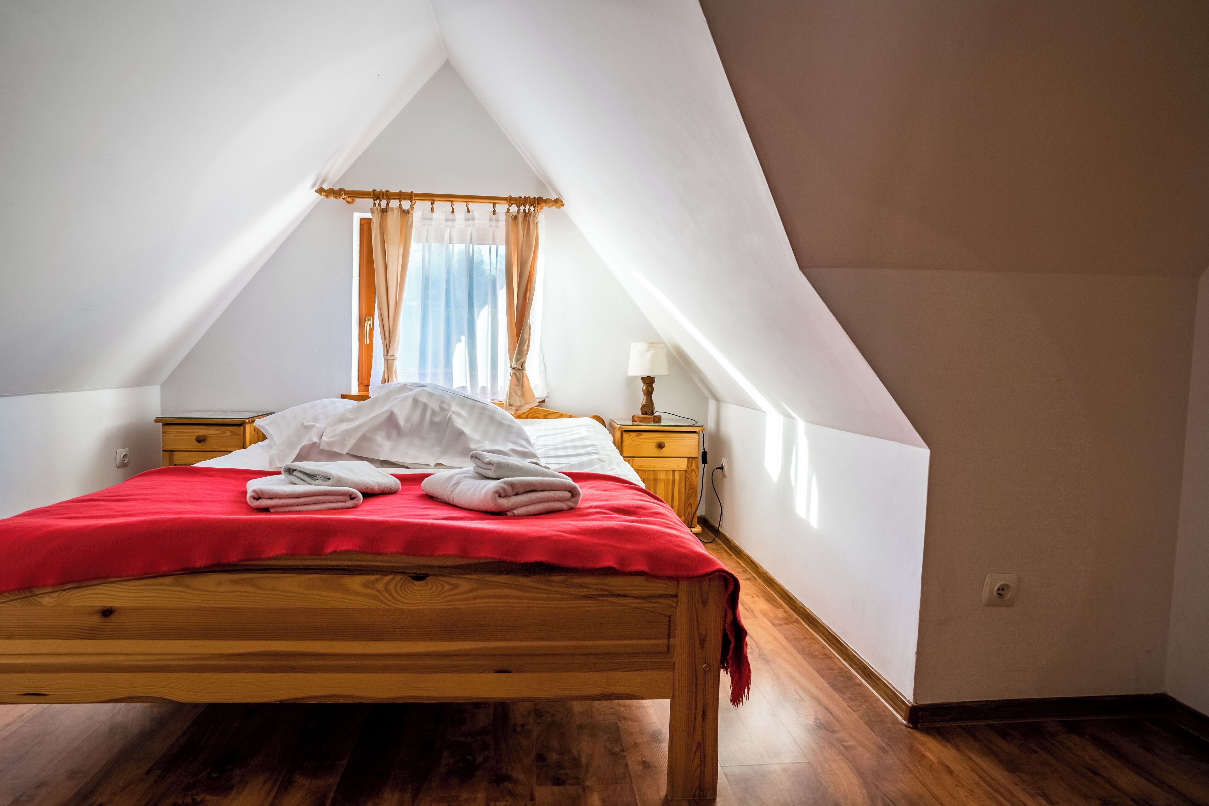 Un dormitorio con cama de madera, ropa de cama roja, una mesita de noche con lámpara y una ventana con cortinas.