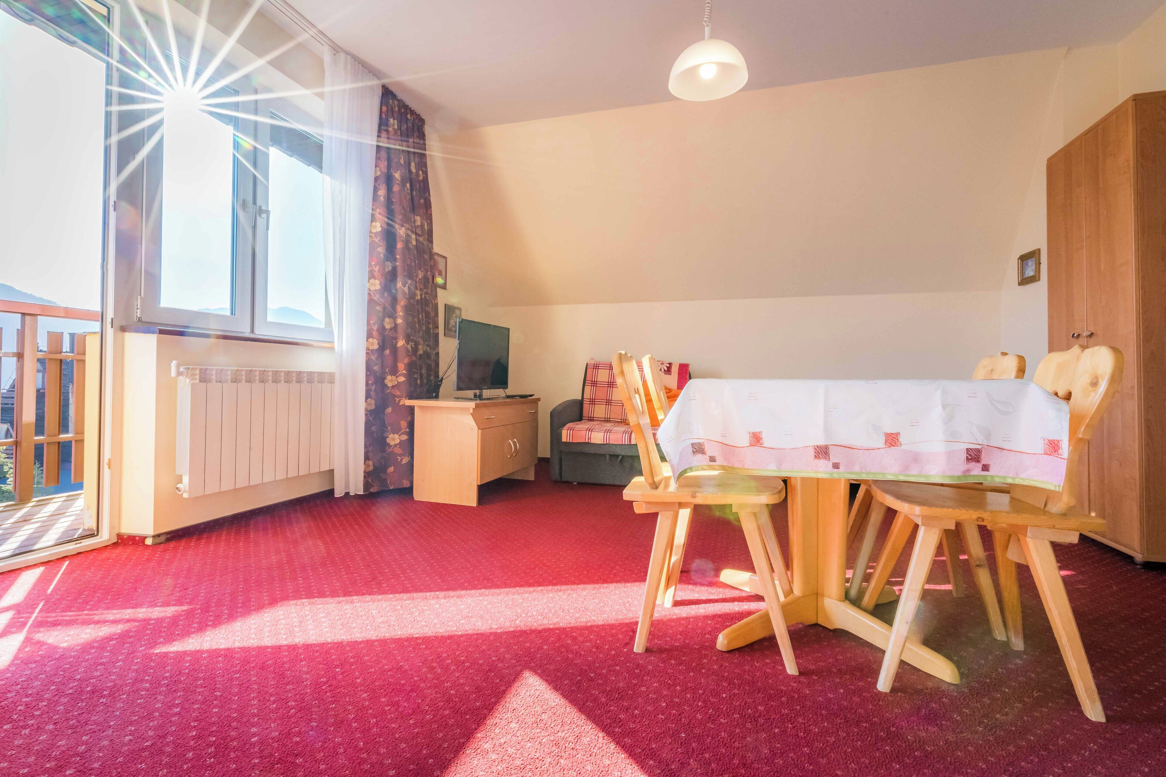 A room with a red carpet, a dining table with chairs, a television, and a wardrobe.
