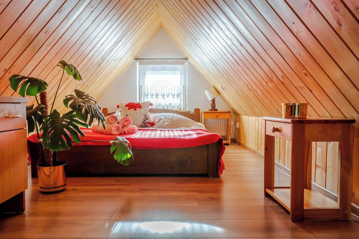 Ett mysigt sovrum på vinden med ett sluttande tak, en säng med ett rött överkast, ett sängbord med en lampa och en krukväxt.