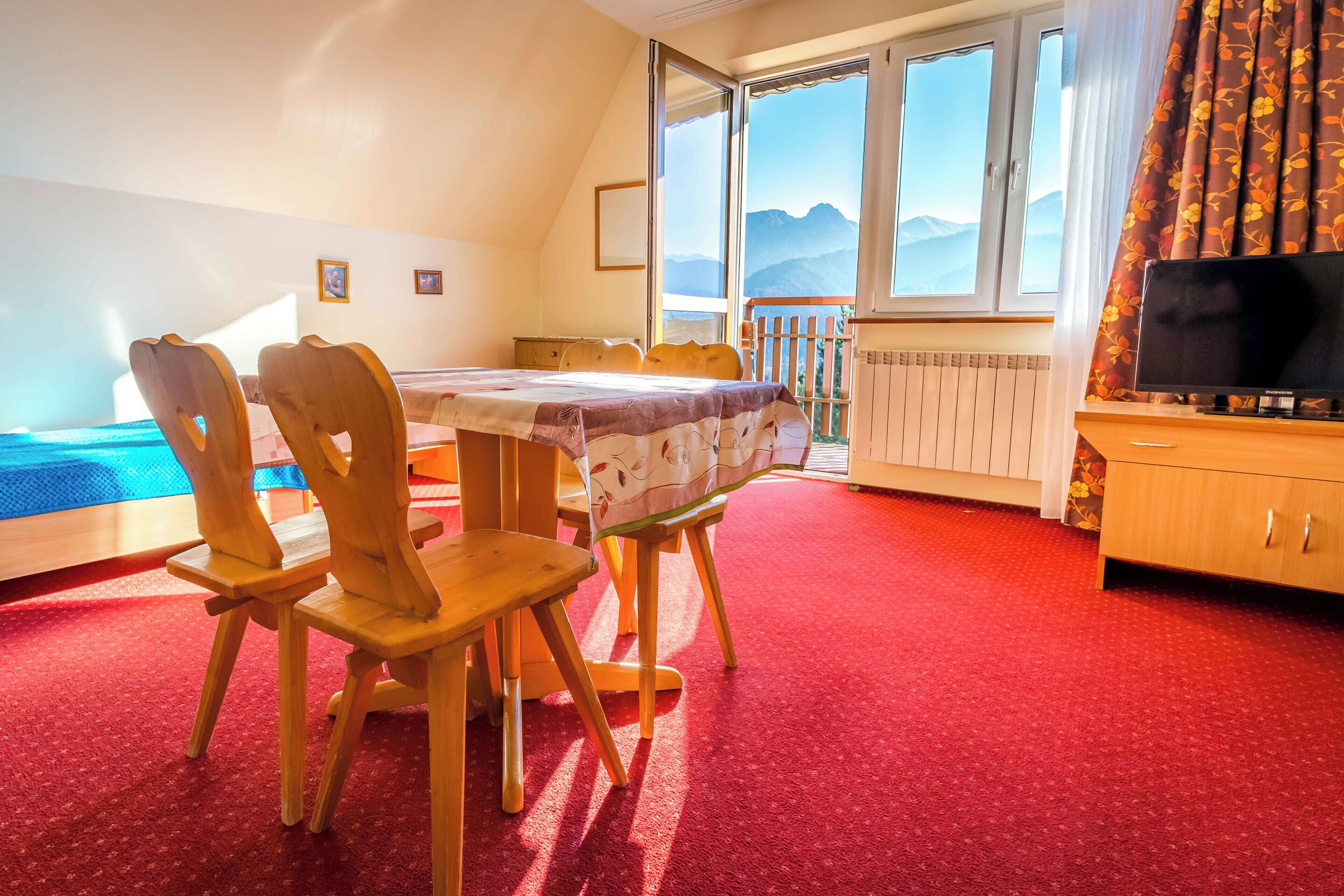 A room with a dining table, chairs, a TV, and a view of mountains through large windows.